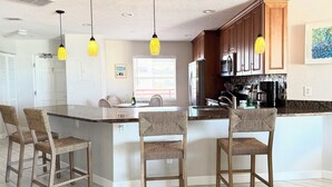 A kitchen with a central island, bar stools, a refrigerator, microwave, and a window with blinds. - Upscale Beachfront Condo • Gulf View Balcony • Pool• Steps to Johns Pass &Dining (Madeira Beach)