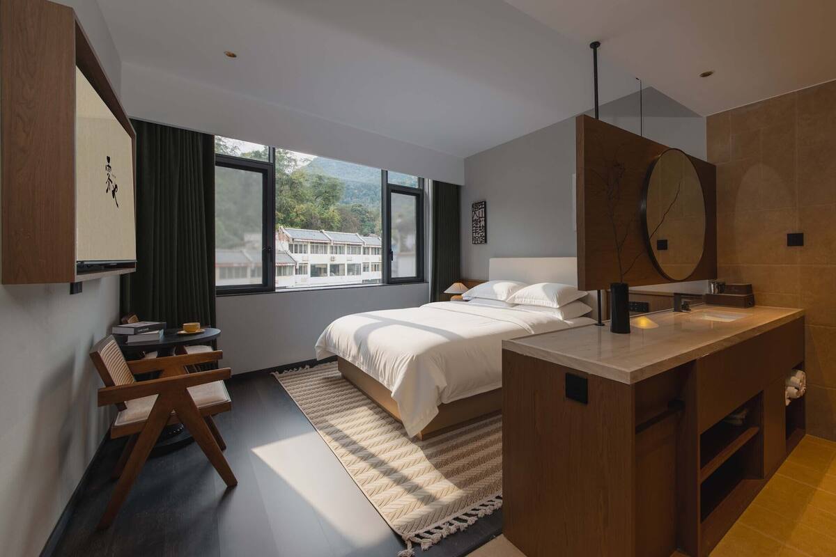 A modern hotel room with a large bed, a desk, a vanity with a mirror, and a view of traditional buildings through the window.