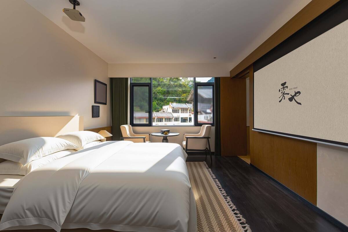 A hotel room with a bed, two chairs, a small table, and a large window with a view of buildings.