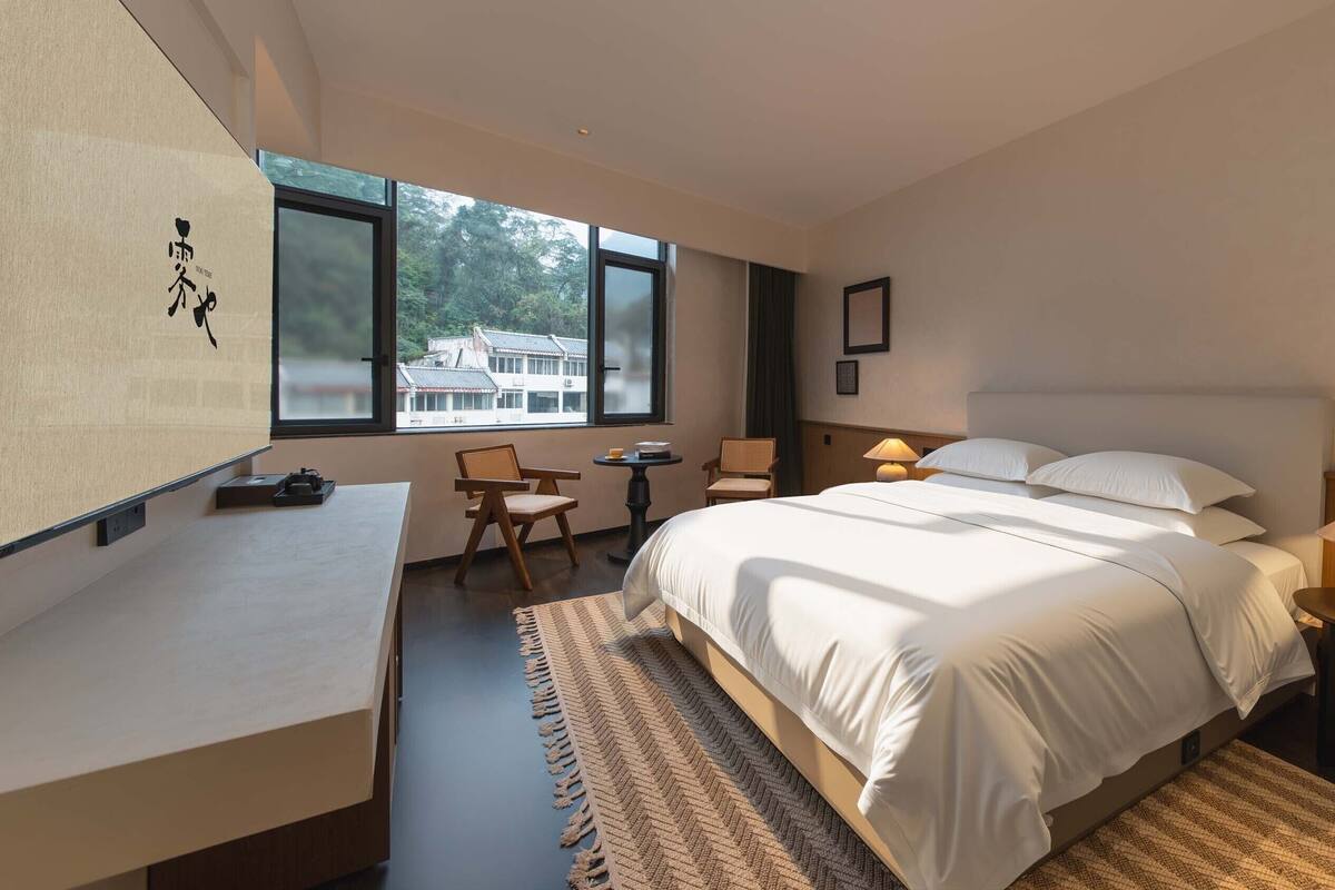 A modern hotel room with a large bed, a desk, a chair, and a view of buildings outside the window.