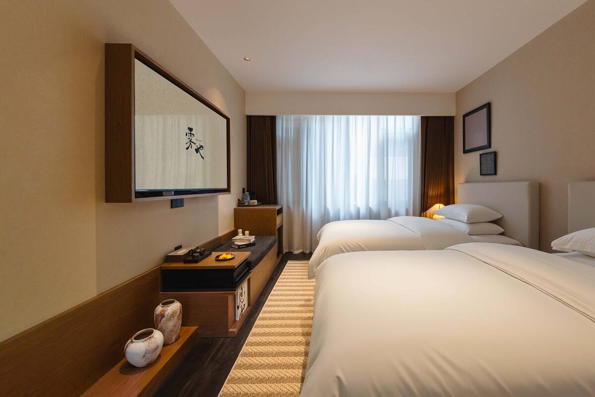 A hotel room with two beds, a desk with a computer, a flat-screen TV, and a large window with curtains.
