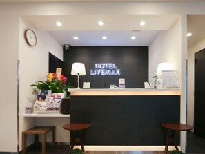 A hotel reception desk with a sign reading 'HOTEL LIVEMAX,' two small tables, a clock, and a plant. - Hotel Livemax Yokohama Station West Exit (Yokohama)