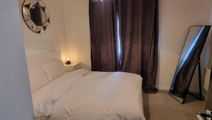 A hotel room with a bed, a nightstand with a lamp, a mirror, and a large window with curtains. - East London 2-Bed Ap Sleeps 4 near Hainault Forest (Romford)