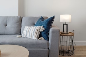 Un sofá gris con un cojín a rayas y un cojín azul, una mesita redonda de madera con una lámpara, y un taburete de estructura metálica negra.