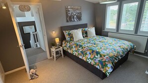 Une chambre à coucher comprenant un lit, une table de chevet, une lampe, une fenêtre avec des stores et une porte donnant sur une autre pièce.