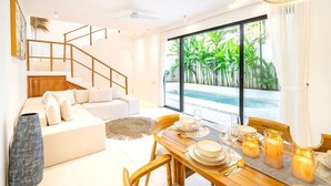 A modern living room with a sofa, dining table set, staircase, and a view of a pool and greenery. - Casa Myla Villa - Bingin Bali (Munggu)