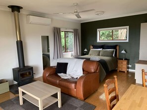 A room with a bed, a sofa, a table, a wooden chair, a heater, a ceiling fan, a window, and a nightstand.
