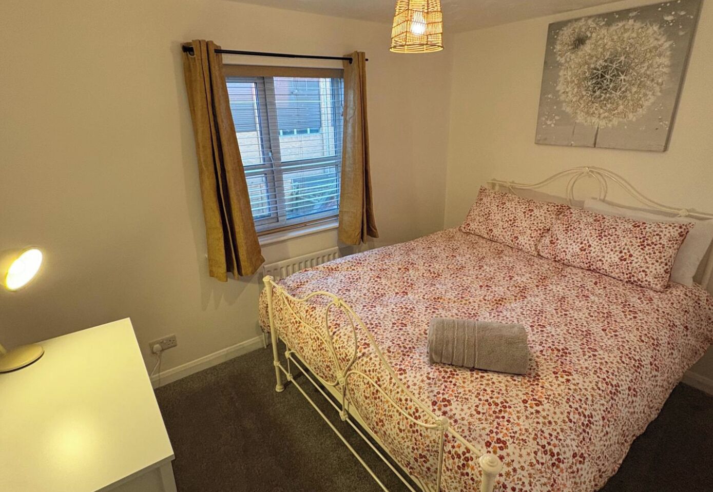 A bedroom with a bed, a window with curtains, a radiator, a lamp, and a wall art piece.