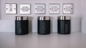 Tre cylindriska behållare märkta med "TE", "KAFFE" och "SOCKER" står på en vit yta, och ovanför dem finns en rad ljusströmbrytare.