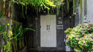 Una entrada estrecha con una puerta blanca que indica '3A Gypsy Villa', rodeada de abundante follaje y plantas.