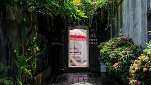 Un estrecho callejón con una hamaca y una cama visibles a través de un vano de puerta, rodeado de exuberante vegetación y un letrero que dice 'GYPSY VILLA'.