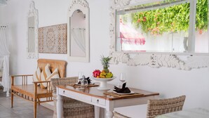 Un comedor con mesa de madera, sillas de mimbre y pared blanca con espejos decorativos y una ventana con plantas.