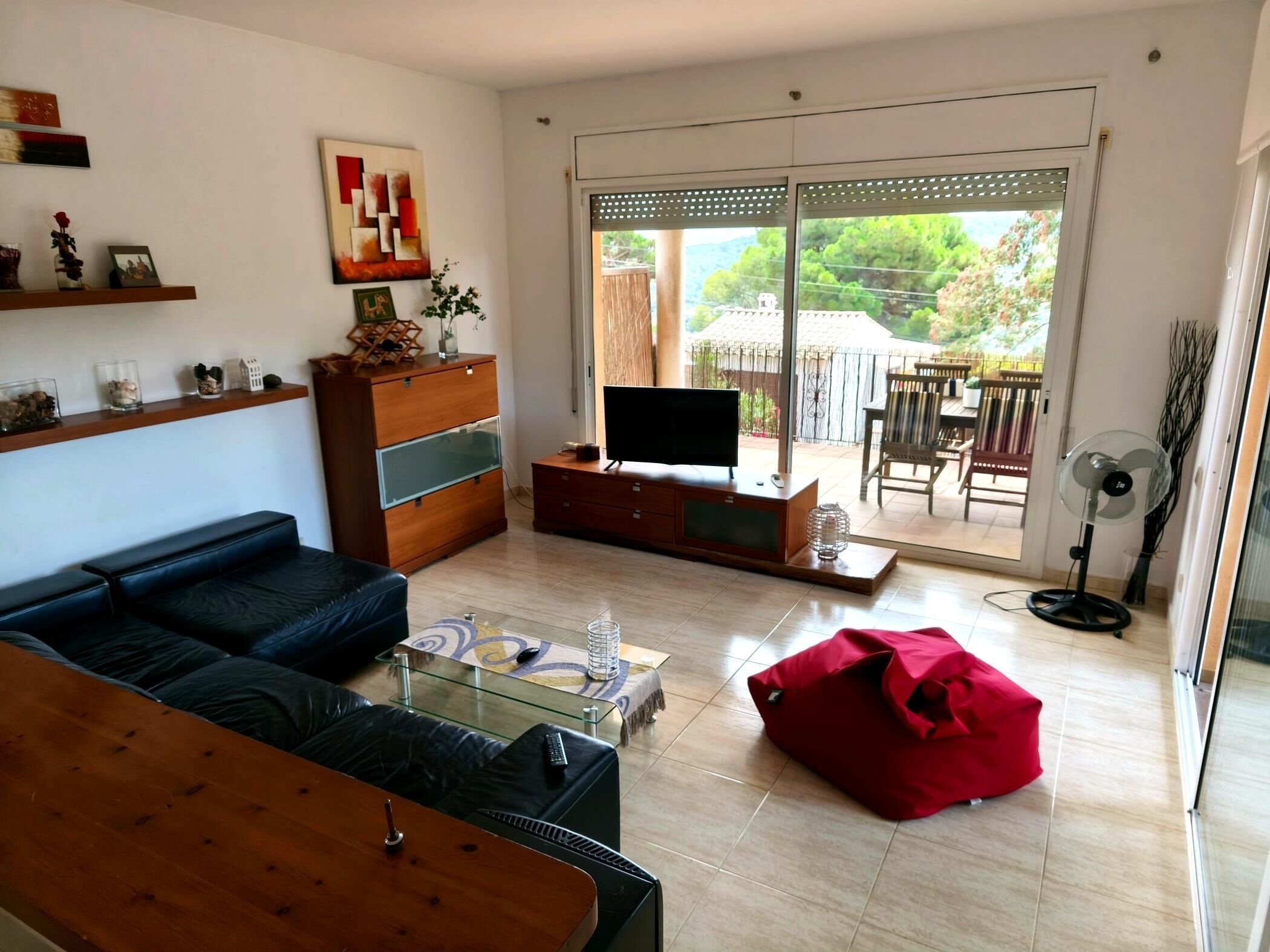Een woonkamer met een bank, televisie, glazen salontafel, rode kruikzitkussen, houten kast en een schuifpui naar een buitenruimte.