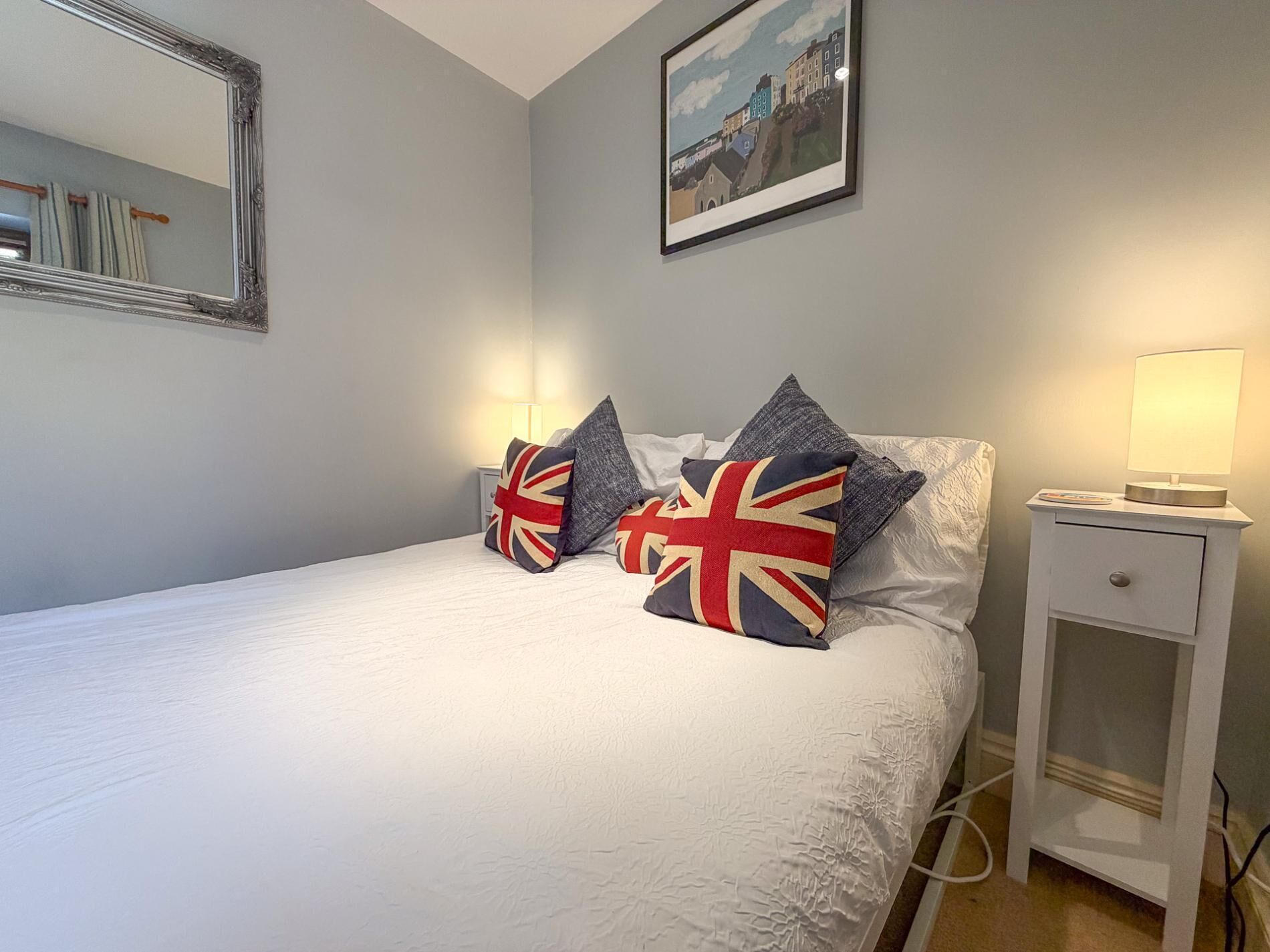Een netjes opgemaakt bed met kussens, een nachtkastje met een lamp, een lijst met een afbeelding aan de muur en een spiegel.
