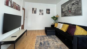 A living room with a flat-screen TV, a black sofa with yellow and purple cushions, a wooden floor, and a large cityscape picture on the wall. - Tottenham - Sleeps 10 - Garden - Near Transport (London)