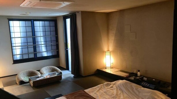 A hotel room with a bed, a small seating area, a window with blinds, a nightstand, and a lamp. - GOOD HOTEL OSAKA (Toyonaka)