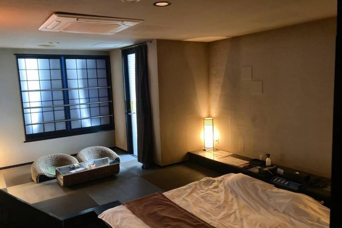 Habitación de hotel con cama, un pequeño espacio para sentarse, ventana con persianas, mesita de noche y lámpara.