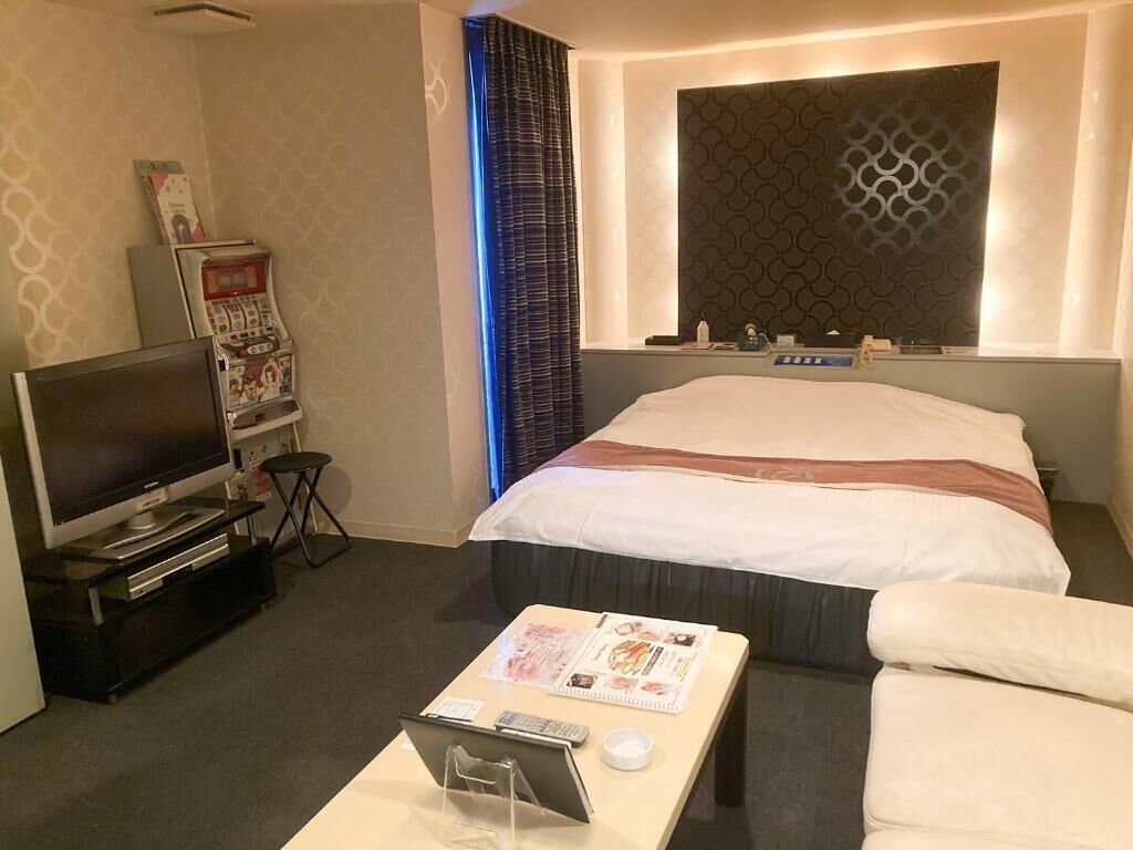 A hotel room with a bed, a TV, a desk with a remote, a magazine, and a wall-mounted control panel.