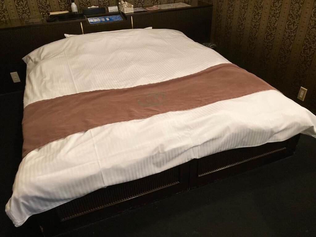Un lit recouvert d’une couverture blanche et marron, avec une tête de lit sombre et une table de chevet en bois sur laquelle se trouvent une lampe et une bouteille.