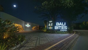 Vista nocturna de la entrada del Complejo Bali Bites, con señalización iluminada, un camino pavimentado y un árbol grande.