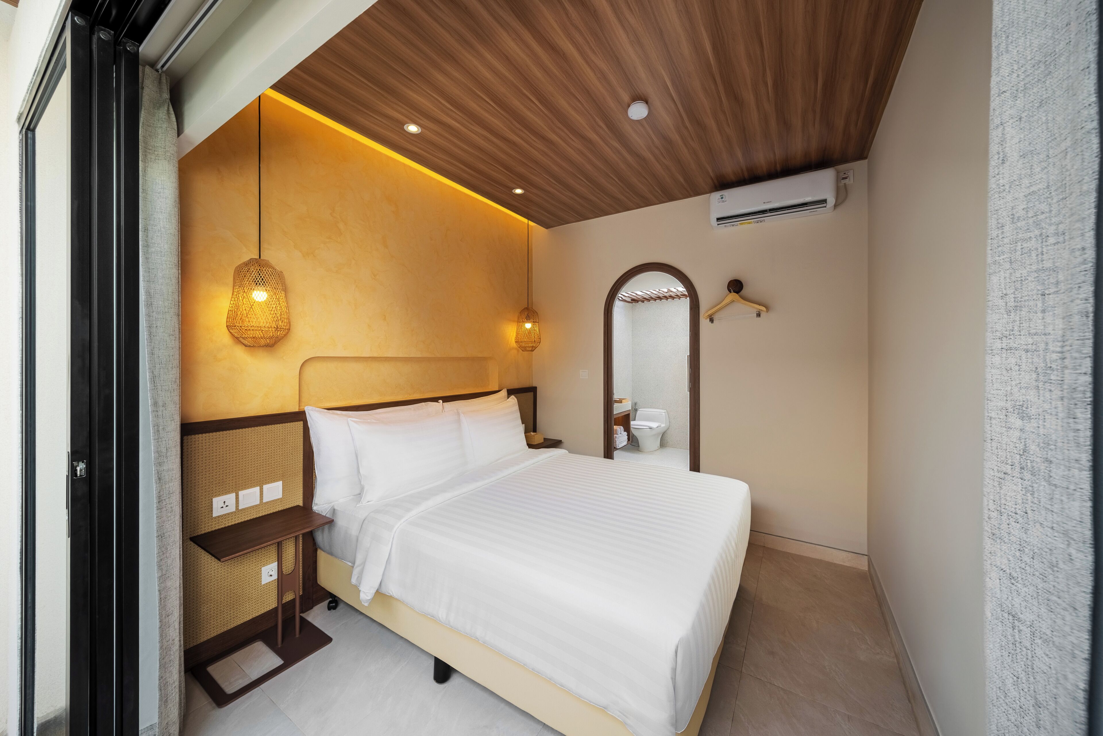 A hotel room with a bed, bedside table, hanging light fixture, and a mirror.