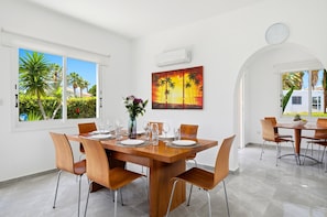 A dining area with a wooden table set for four, a vase with flowers, and a sunset painting on the wall. - Villa Haven by Ezoria Villas (Paphos)