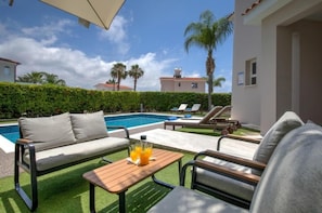 A poolside area with a sofa, lounge chairs, and a table with drinks. - Villa Haven by Ezoria Villas (Paphos)