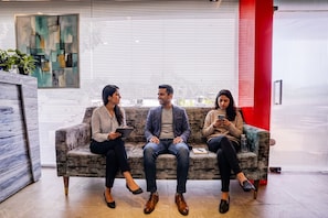 Three individuals seated on a sofa in a modern interior space with a large window, a red accent wall, and a piece of abstract art on the wall. - FabHotel Pride Suitz (Mumbai)