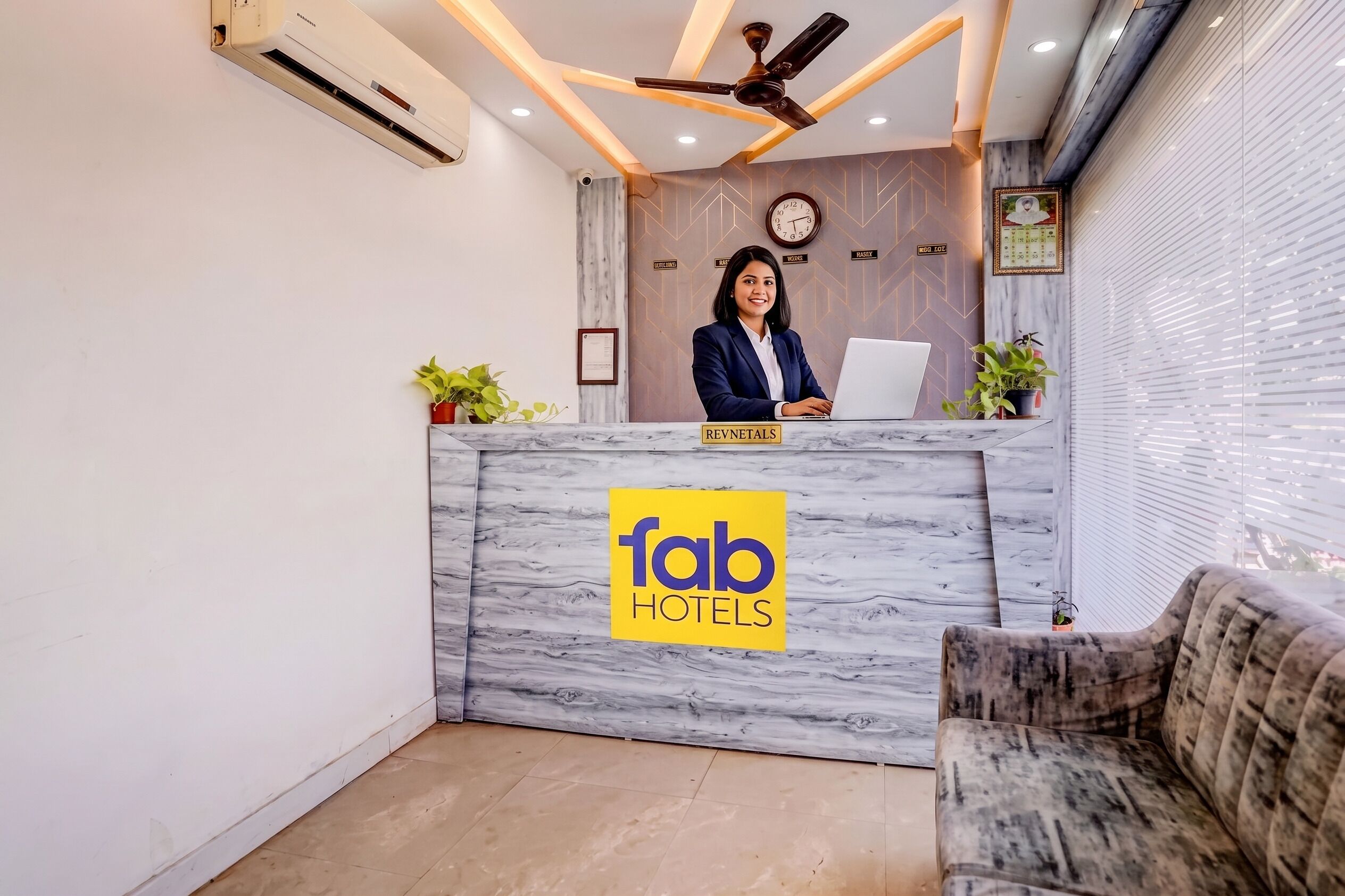 Un comptoir d’accueil avec un plateau en marbre et le logo de « fab hotels ». Un ventilateur de plafond est installé au-dessus, une horloge est accrochée au mur, et un certificat encadré est également visible sur le mur.