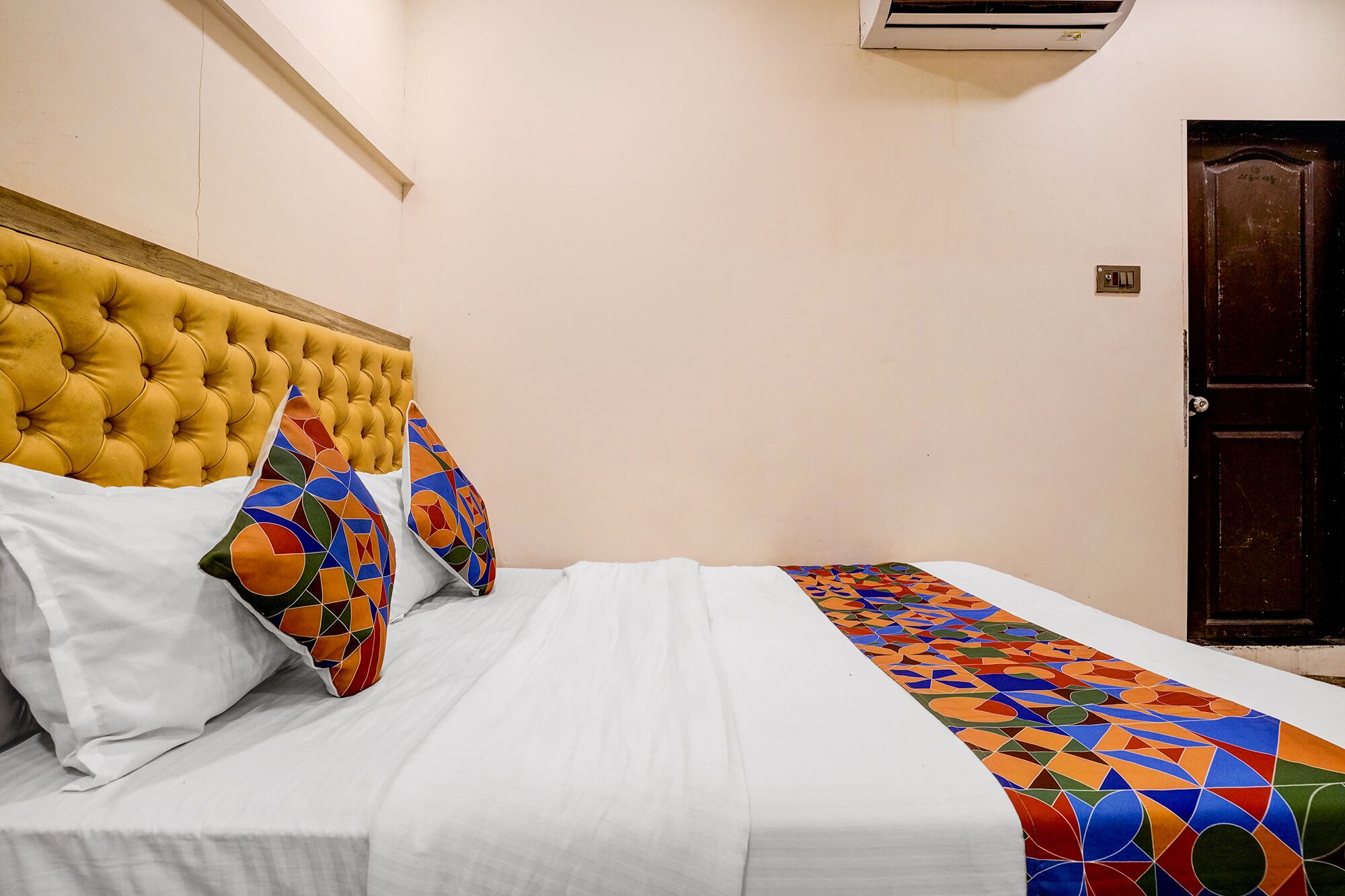 Ein Schlafzimmer mit einem geblatteten Kopfteil, weißer Bettwäsche, bunten Kissen und einer Decke mit geometrischem Muster.
