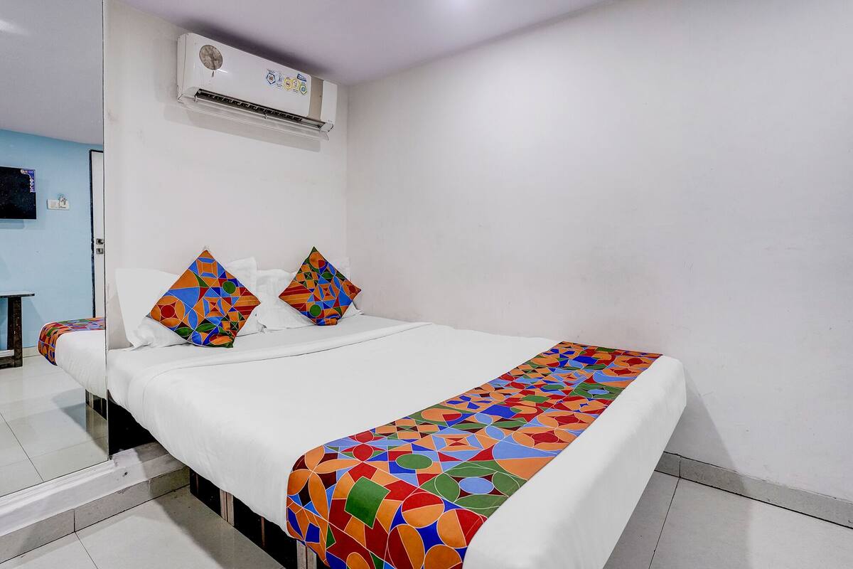 A hotel room with a bed, two pillows, a white wall, an air conditioning unit, and a tiled floor.