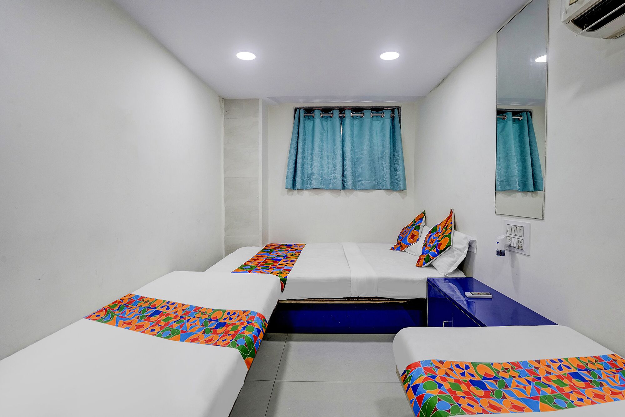 A small, clean room with two beds, a mirror, a window with curtains, and a desk with a chair.