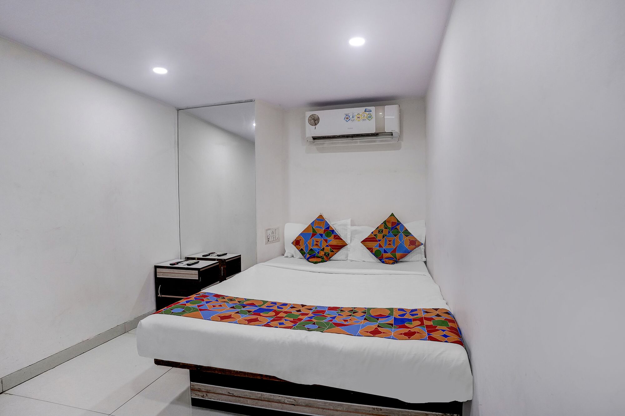 A bedroom with a bed, a bedside table, an air conditioning unit, and a wall-mounted mirror.