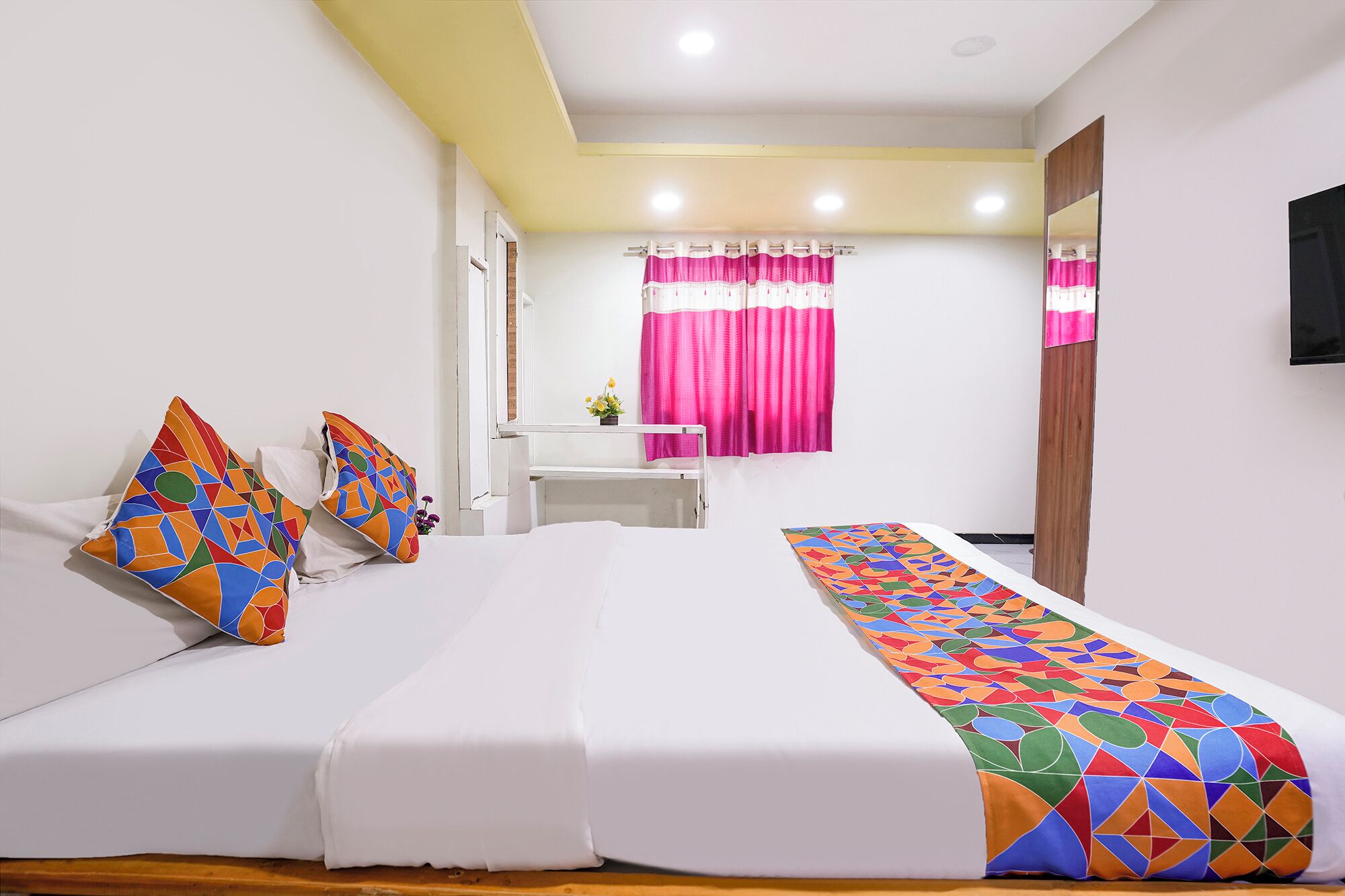 A hotel room with a bed, pillows, a window with curtains, a mirror, and a television.