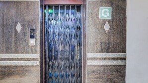 Een metalen liftdeur met een ruitpatroon, een groen ‘G’ bordje en een klein bedieningspaneel.