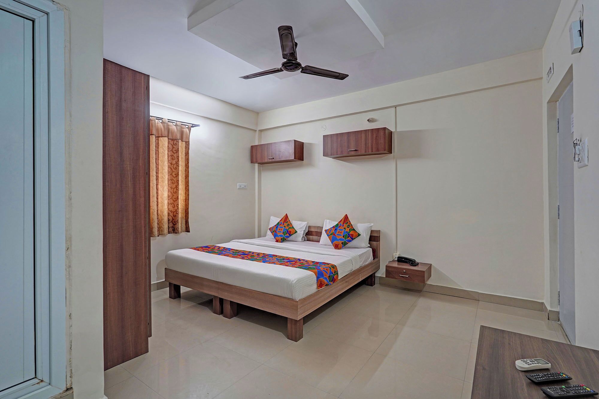 Une chambre à coucher comprenant un lit, un ventilateur de plafond, une armoire en bois et une petite table avec une télécommande.