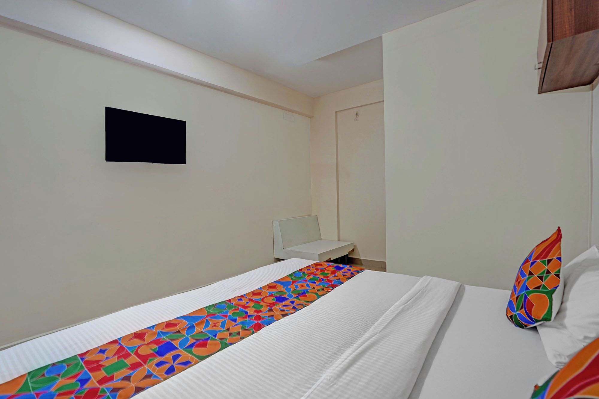 A hotel room with a bed, a small bedside table, a wall-mounted TV, and a colorful pillow.