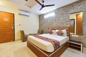 A bedroom with a bed, a chair, a nightstand, a ceiling fan, a wall-mounted mirror, and a wooden door. - FabHotel Prime Royal Elite - Nr Jaipur International Airport (Jaipur)