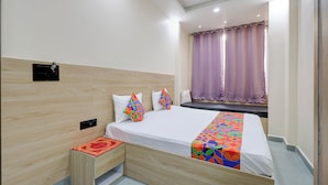 Habitación de hotel con cabecera de madera, cama con ropa de cama colorida, mesita de noche con un tapete rojo y ventana con cortinas moradas.