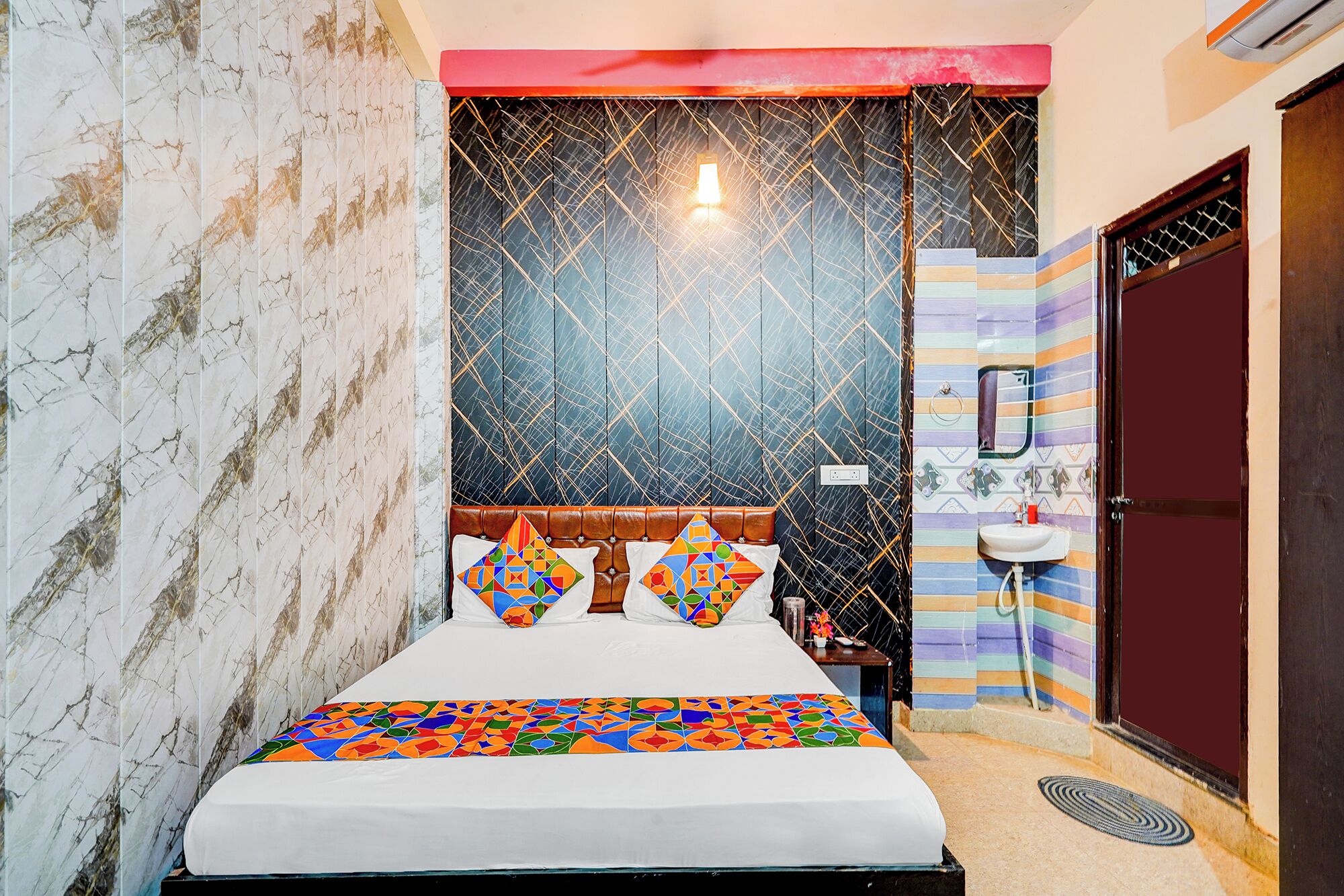 A bed with a colorful geometric bedspread, a headboard with a light fixture, and a striped wall with a sink and mirror.