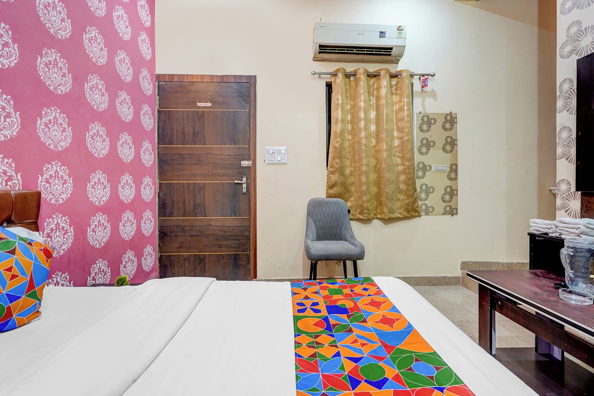 A room with a bed, a chair, an air conditioning unit, a curtain, and a table with a water bottle.