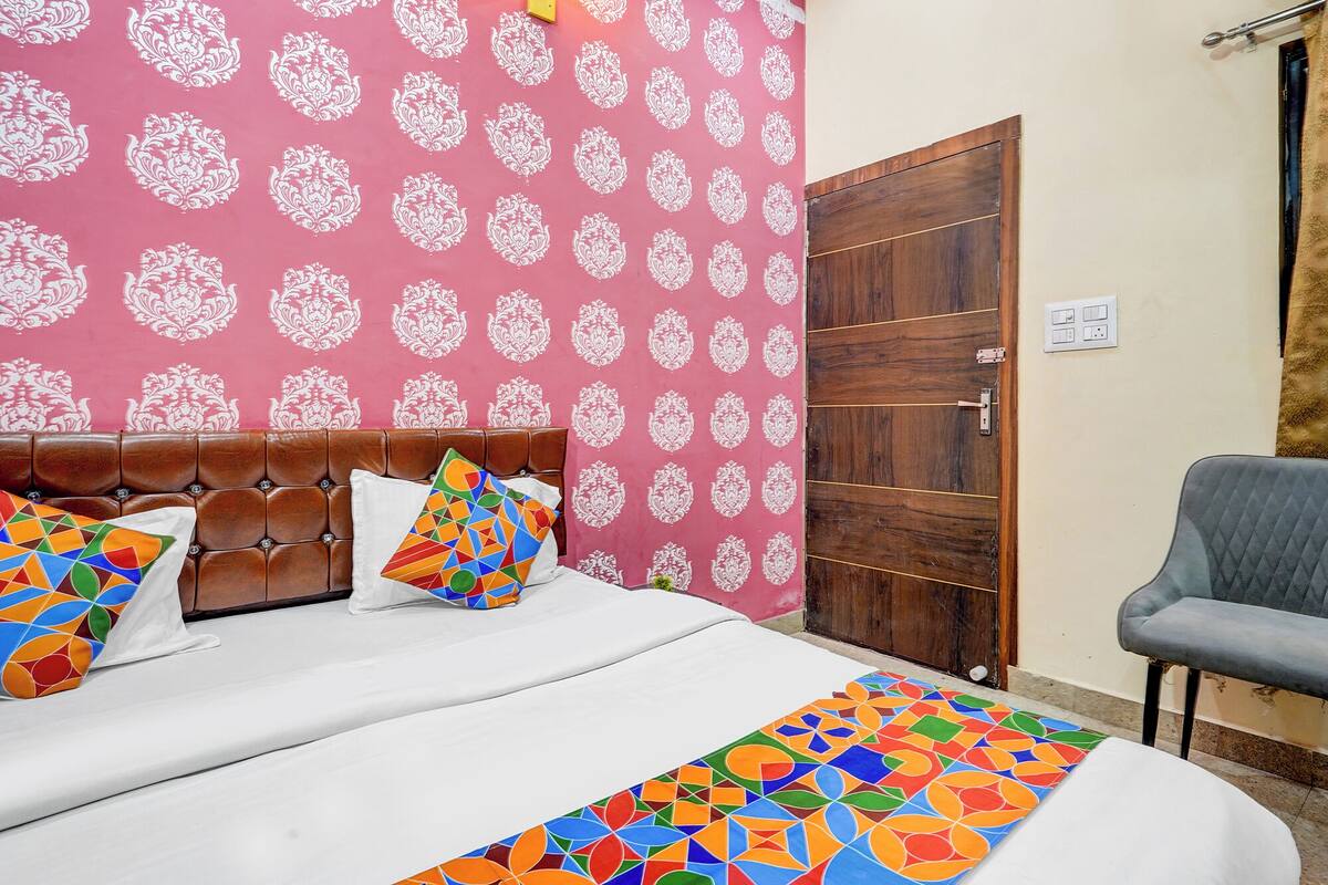 A bedroom with a patterned wallpaper, a brown upholstered headboard, a bed with white bedding, colorful pillows, a wooden door, and a grey chair.