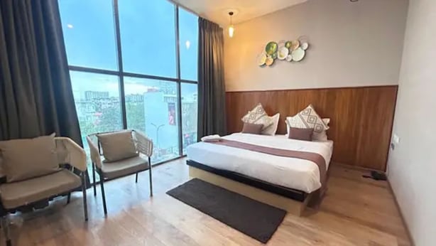 Ett hotellrum med en stor säng, en stol, utsikt genom ett stort fönster och en väggmonterad lampa.