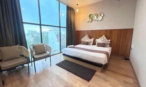 A hotel room with a large bed, a chair, a view through a large window, and a wall-mounted light fixture. - Fabhotel Kg Estate (Jaipur)