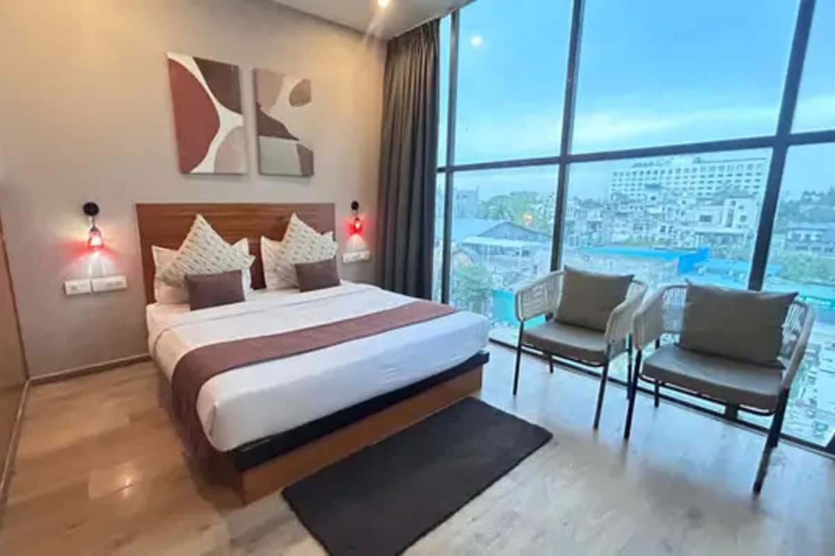 A modern hotel room with a large bed, two bedside lamps, a view of the city through large windows, and a seating area with two chairs.