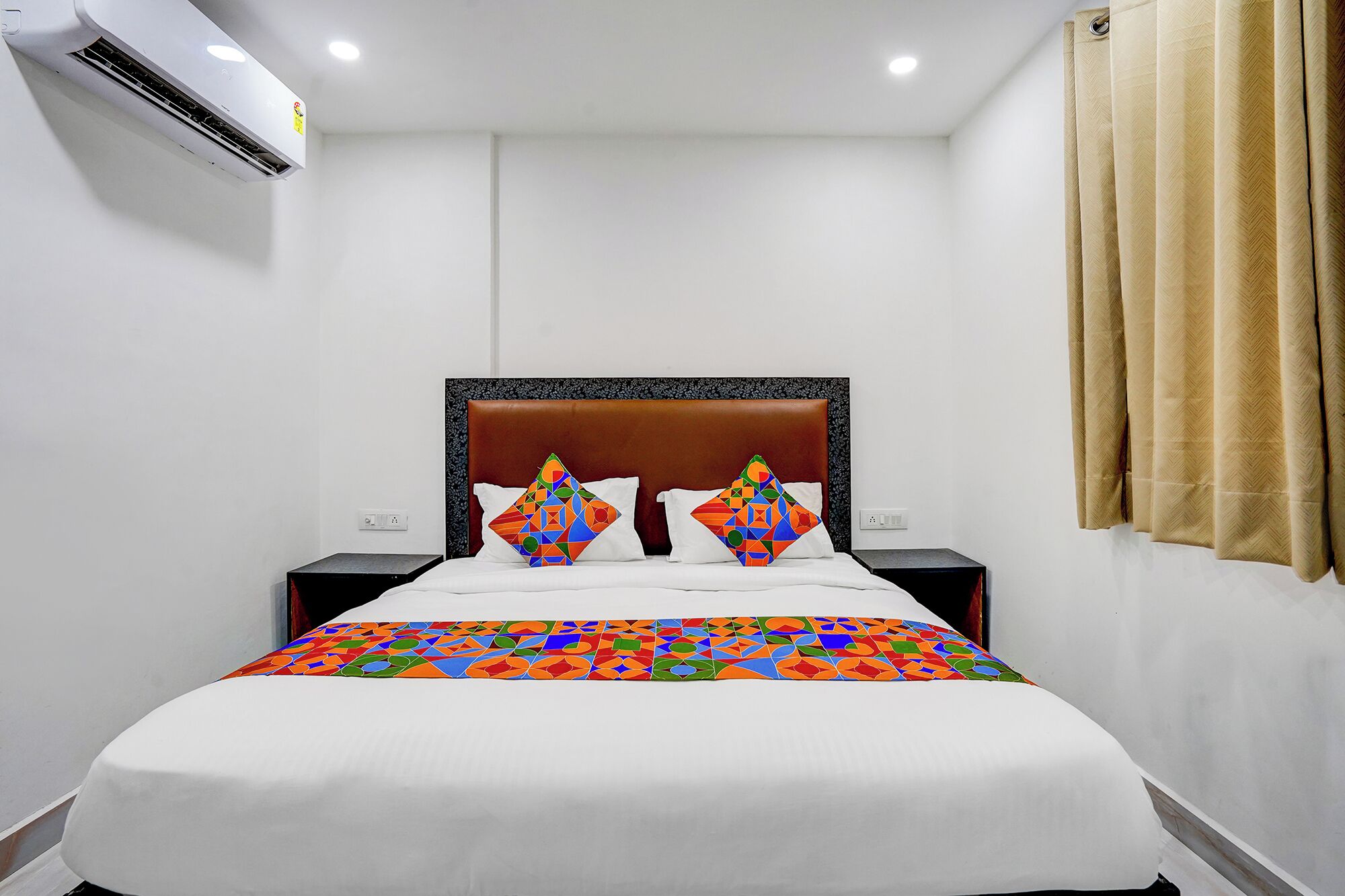 A hotel room with a bed, two nightstands, a brown headboard, a beige curtain, and an air conditioning unit on the wall.