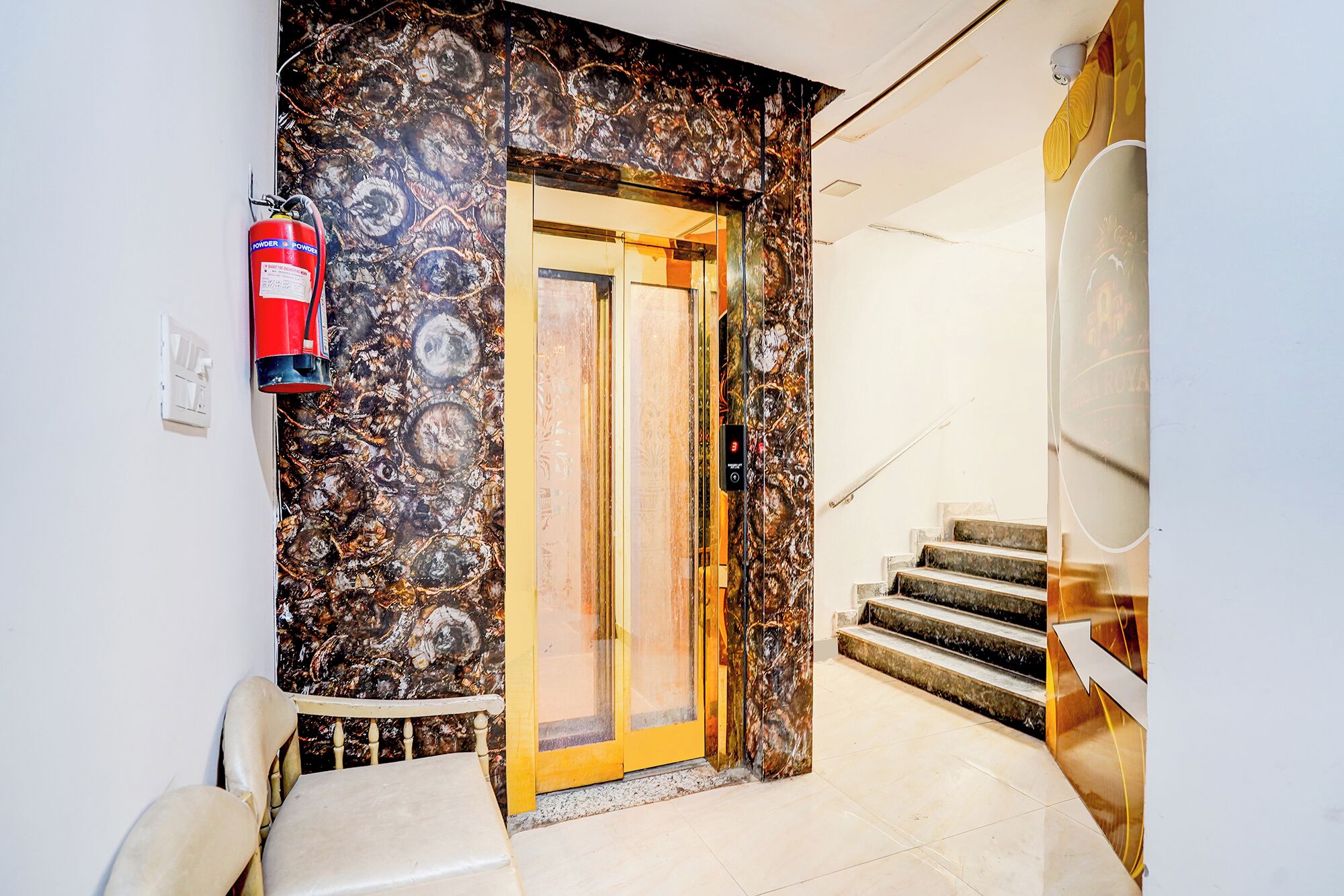A hotel entrance with a marble wall, a fire extinguisher, a staircase, and a door with a digital display.