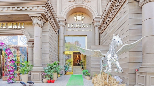 A grand entrance with a horse sculpture, a sign reading 'L'ELEGANT THE ROYAL HOTEL & BANQUETS', and a green carpet leading to the door.