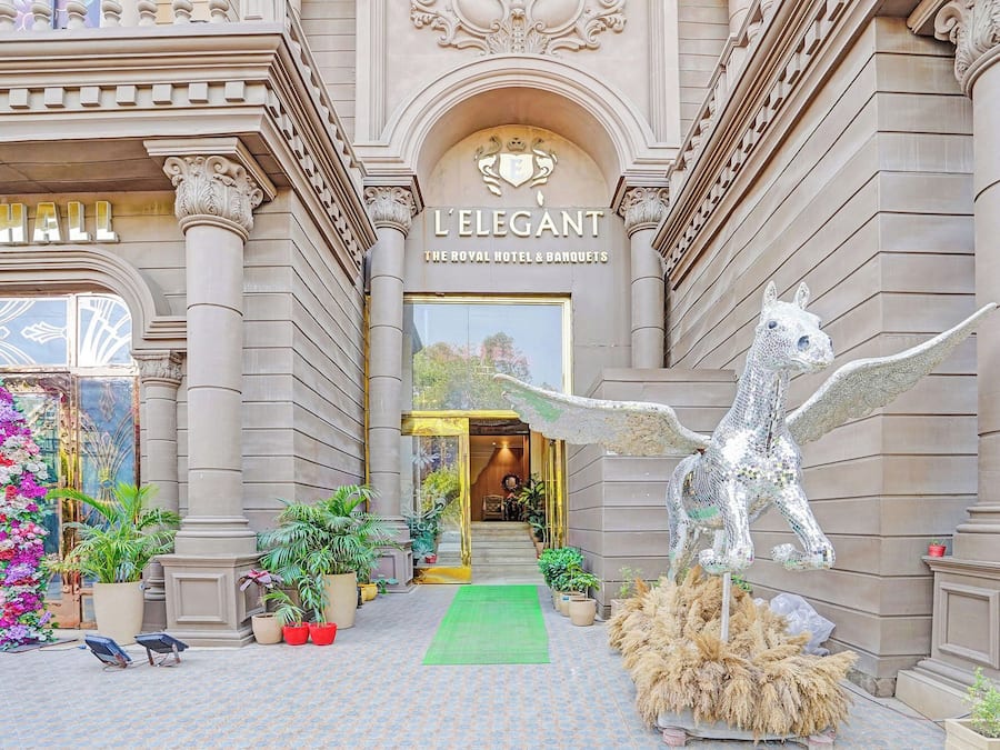 A grand entrance with a horse sculpture, a sign reading 'L'ELEGANT THE ROYAL HOTEL & BANQUETS', and a green carpet leading to the door.
