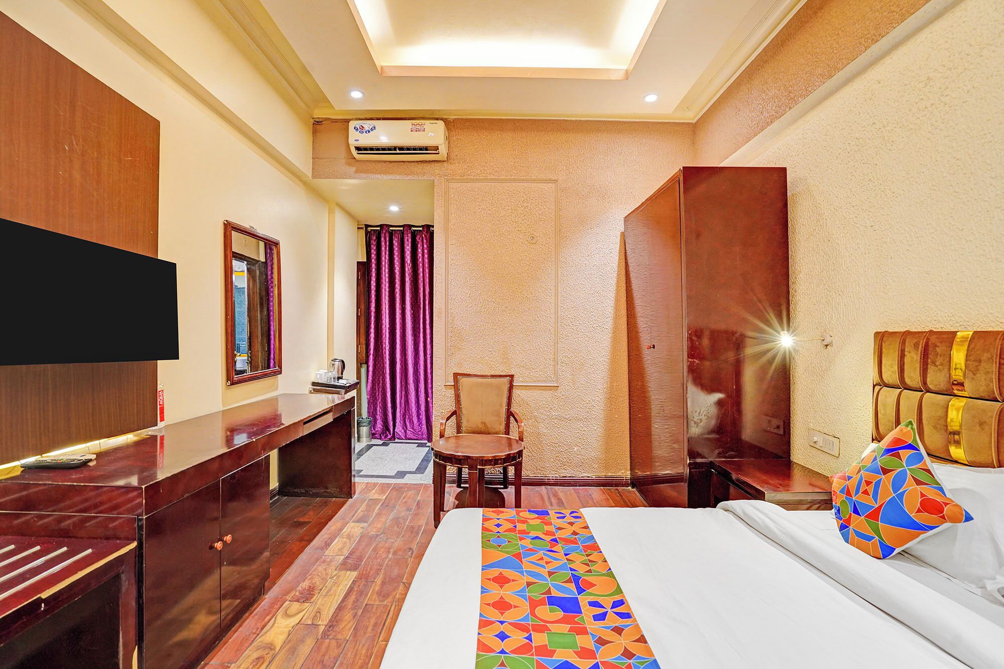 A hotel room with a bed, a desk, a chair, a television, and an air conditioning unit.